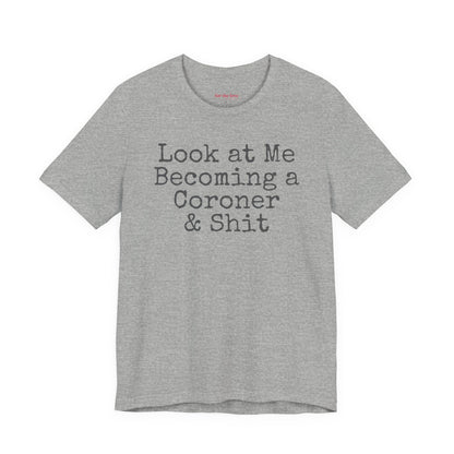 Grey funny coroner T-shirt featuring the phrase Look at Me Becoming a Coroner & Shit in vintage typewriter-style lettering.