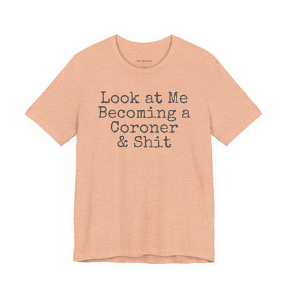 Peach funny coroner T-shirt featuring the phrase Look at Me Becoming a Coroner & Shit in vintage typewriter-style lettering.