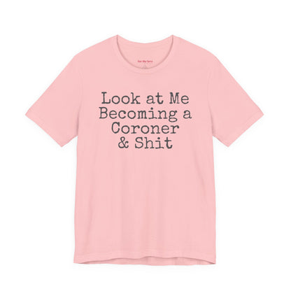 Pink funny coroner T-shirt featuring the phrase Look at Me Becoming a Coroner & Shit in vintage typewriter-style lettering.