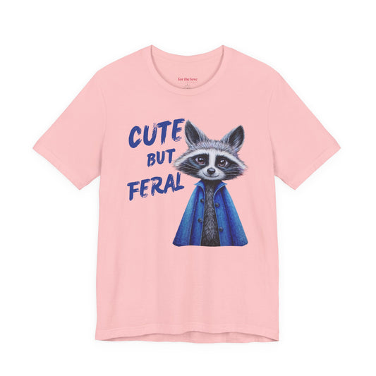 Pink graphic t-shirt featuring a cute raccoon in a blue coat with the phrase “Cute But Feral”