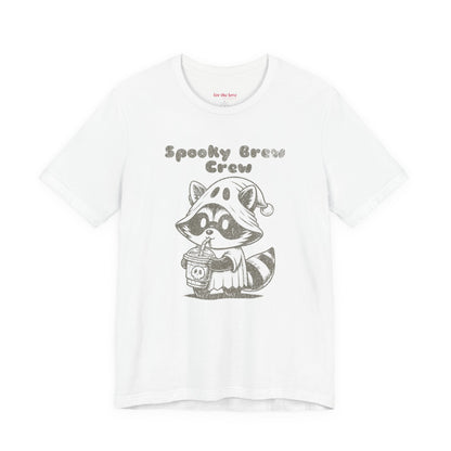 white graphic t-shirt featuring a cute raccoon dressed as a ghost drinking coffee with the words “Spooky Brew Crew”