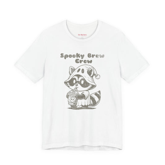 white graphic t-shirt featuring a cute raccoon dressed as a ghost drinking coffee with the words “Spooky Brew Crew”